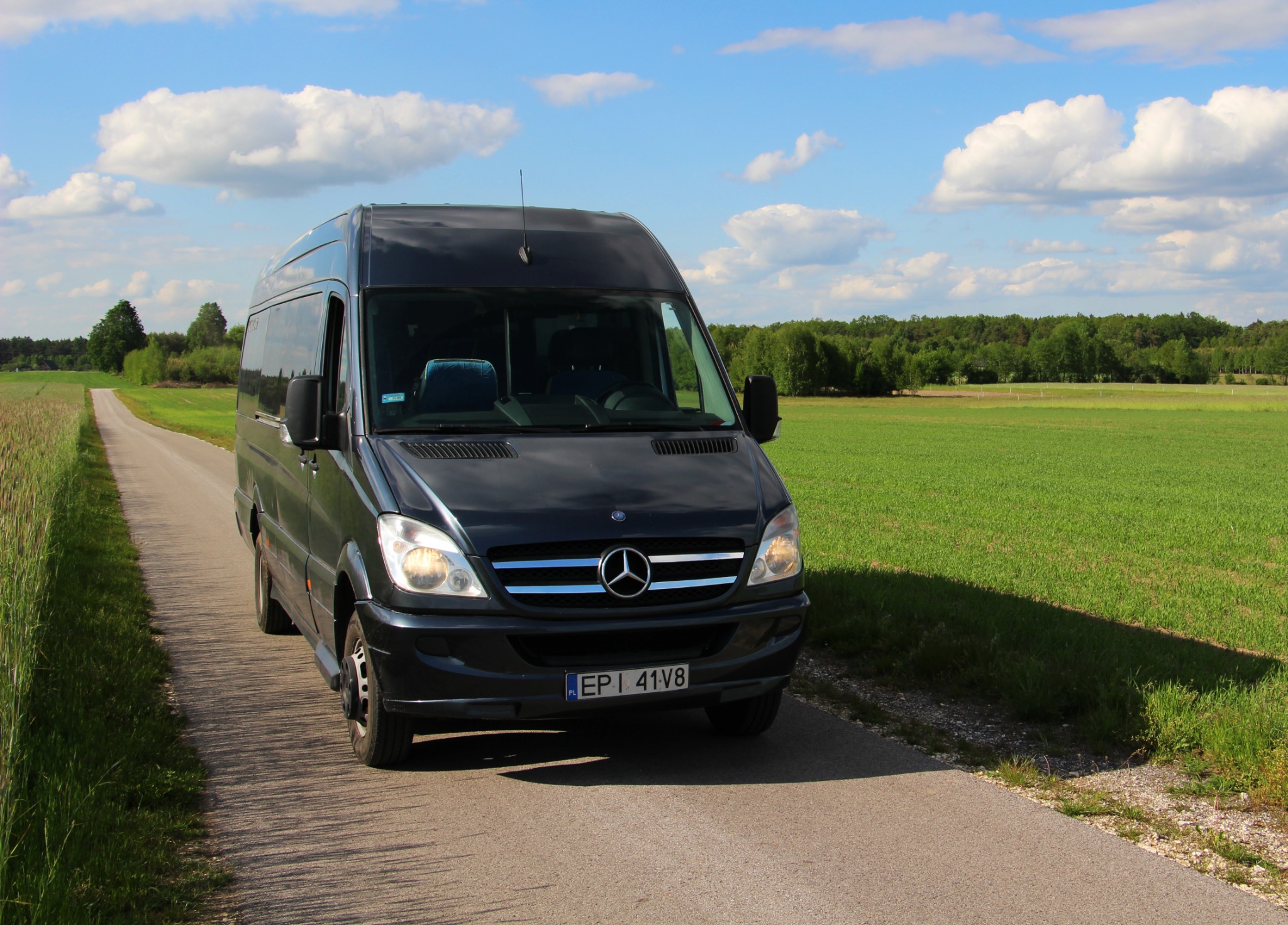 Czarny Mercedes-Benz Sprinter z polską tablicą rejestracyjną EPI 41V8 na wąskiej asfaltowej drodze wśród zielonych pól i lasów w słoneczny dzień.