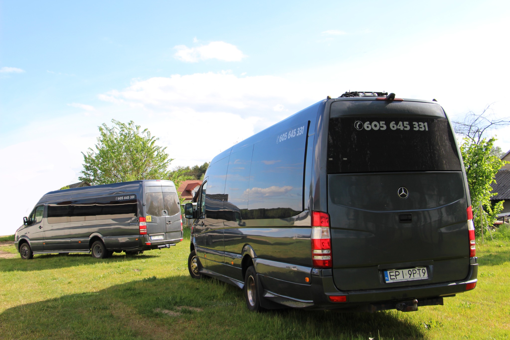 Dwa ciemnoszare busy Mercedes-Benz na trawiastym terenie, tylny bus z numerem telefonu 605 645 331 na szybie i hakiem holowniczym.
