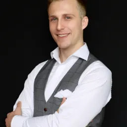 Portret uśmiechniętego brokera ubezpieczeniowego z Lublina w szarej kamizelce w kratę i białej koszuli na czarnym tle.