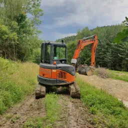 Pomarańczowo-szara mini koparka Hitachi na błotnistej drodze gruntowej, z zielonymi drzewami i krzewami w tle, przygotowana do robót ziemnych.