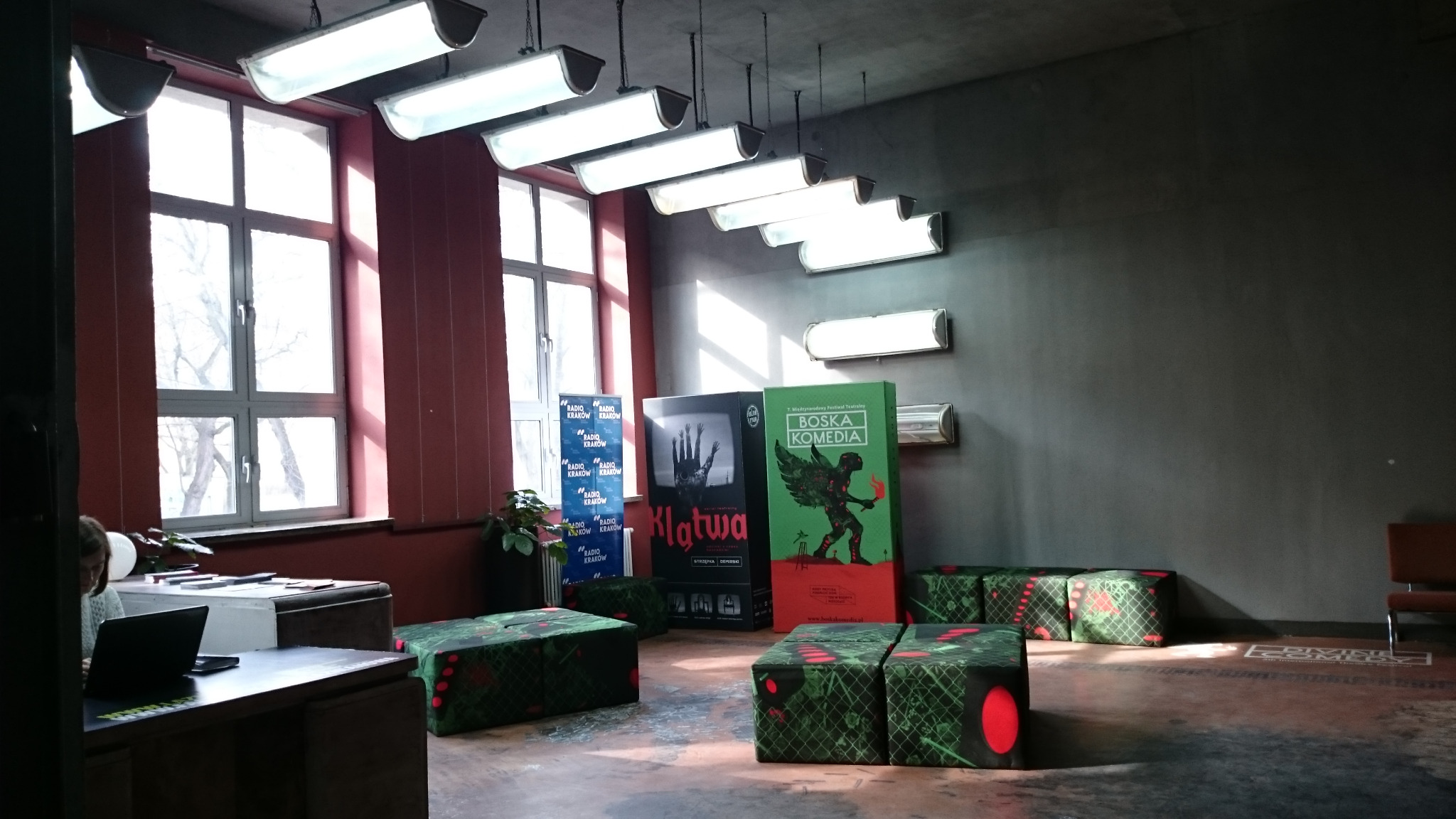 Wnętrze Teatru Łaźnia Nowa z plakatami spektakli 'Klatwa' i 'Boska Komedia', siedziskami w geometryczne wzory i rzędem lamp sufitowych.