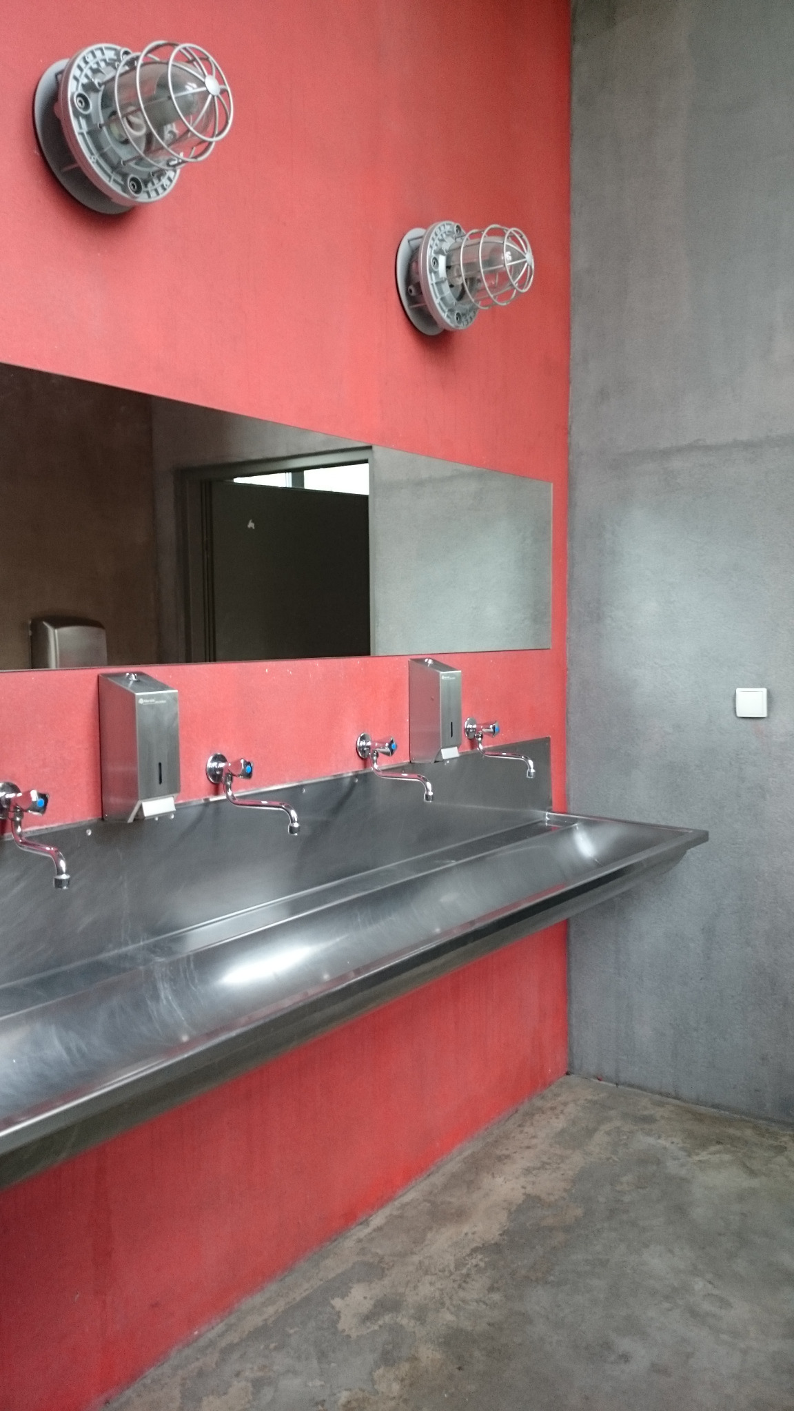 Wnętrze toalety publicznej w Teatrze Łaźnia Nowa: długa, metalowa umywalka z kilkoma kranami i dozownikami mydła, czerwona ściana z dwoma przemysłowymi lampami w metalowych koszach, duże lustro.