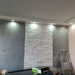 Wykończone wnętrze pokoju z akcentem ściany z białej cegły dekoracyjnej, szara ściana z efektem betonu, podwieszany sufit z oświetleniem punktowym i rozstawiona drabina malarska w rogu.