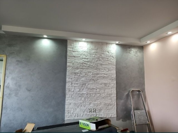 Wykończone wnętrze pokoju z akcentem ściany z białej cegły dekoracyjnej, szara ściana z efektem betonu, podwieszany sufit z oświetleniem punktowym i rozstawiona drabina malarska w rogu.