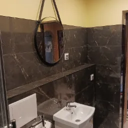 Umywalka z szafką i bidet w wykończonej łazience, ściany wyłożone ciemnymi płytkami imitującymi marmur, okrągłe lustro z czarnym paskiem wiszące na ścianie.