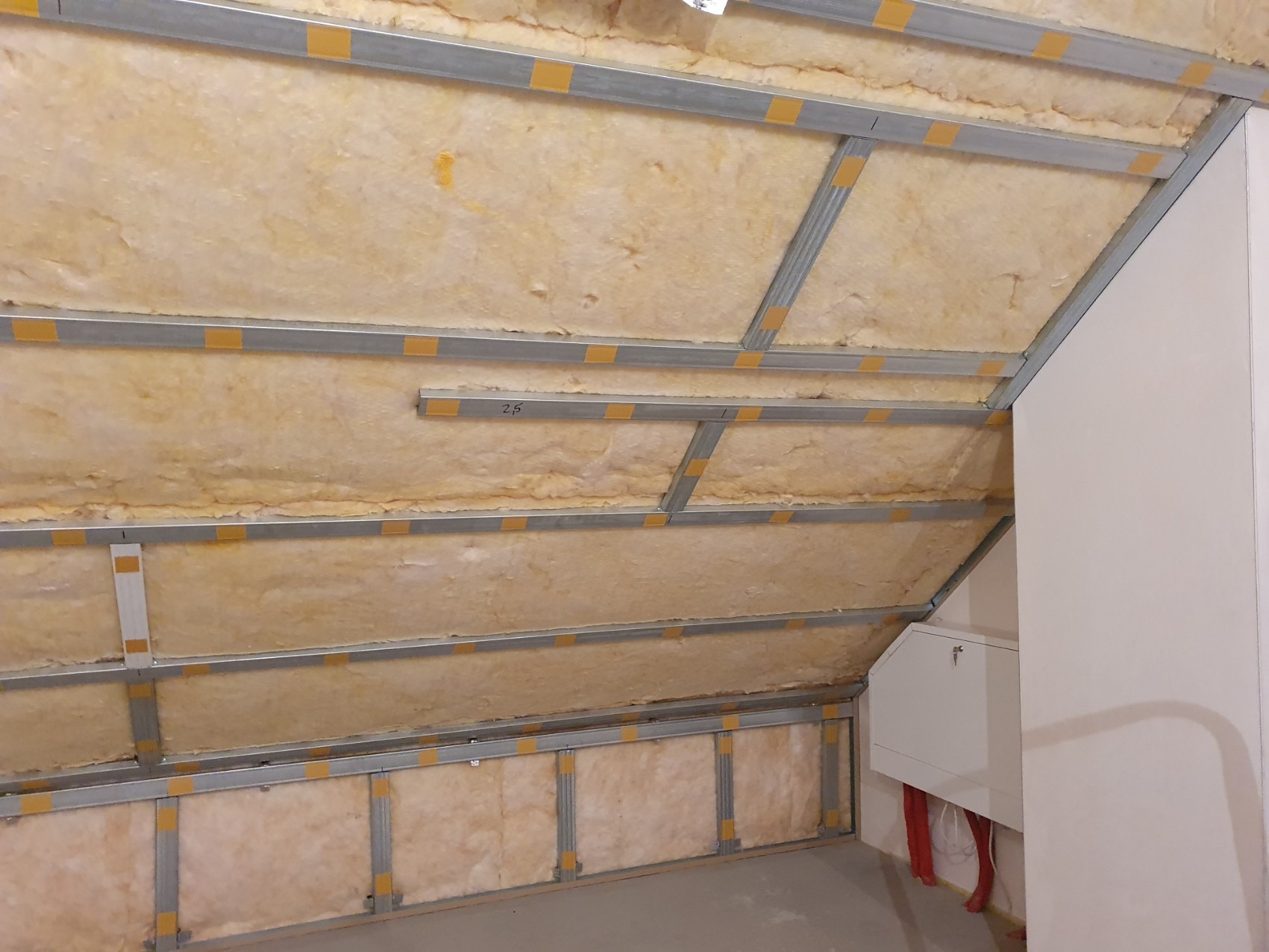 Izolacja poddasza wełną mineralną zamocowaną między metalowymi profilami, widoczne fragmenty instalacji elektrycznej.