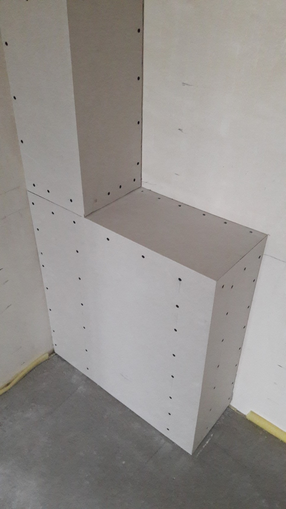 Zabudowa z płyt gipsowo-kartonowych w rogu pomieszczenia, widoczne śruby mocujące, taśma malarska zabezpieczająca podłogę.