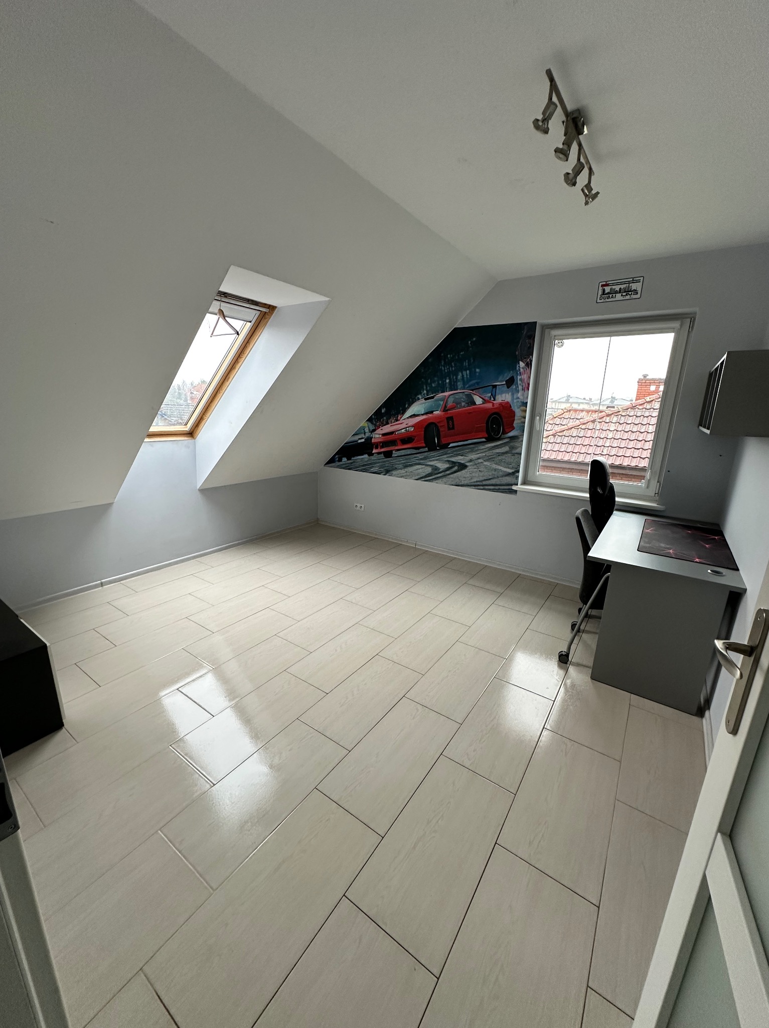 Puste poddasze ze skośnym oknem, jasnymi płytkami podłogowymi, biurkiem komputerowym i tapetą z czerwonym samochodem wyścigowym na ścianie.