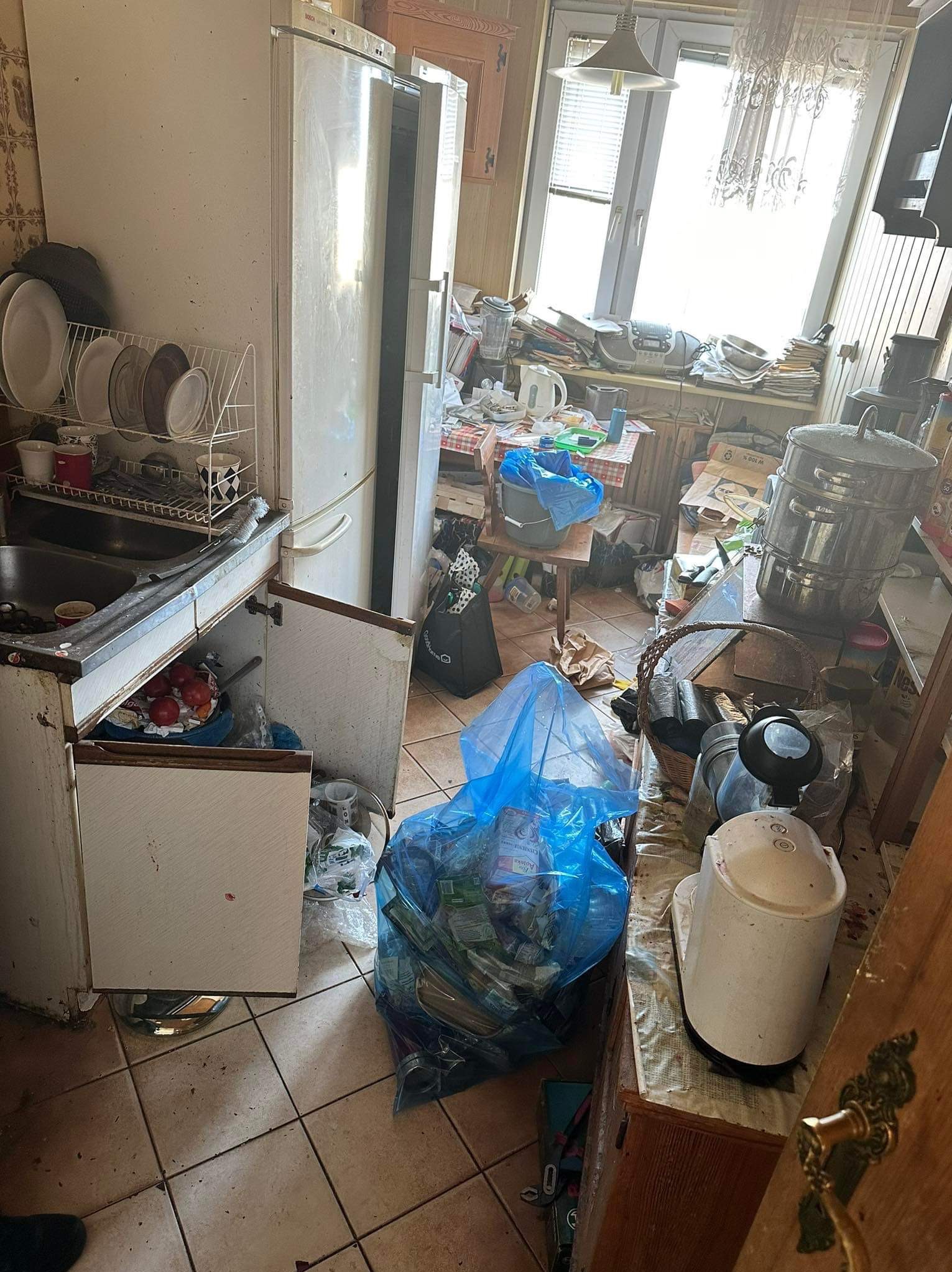 Zaniedbana kuchnia przed sprzątaniem: pełne worki na śmieci, brudne naczynia w suszarce, otwarte szafki z resztkami jedzenia, zagracony blat i stół, widoczne ślady zabrudzeń na podłodze i meblach.