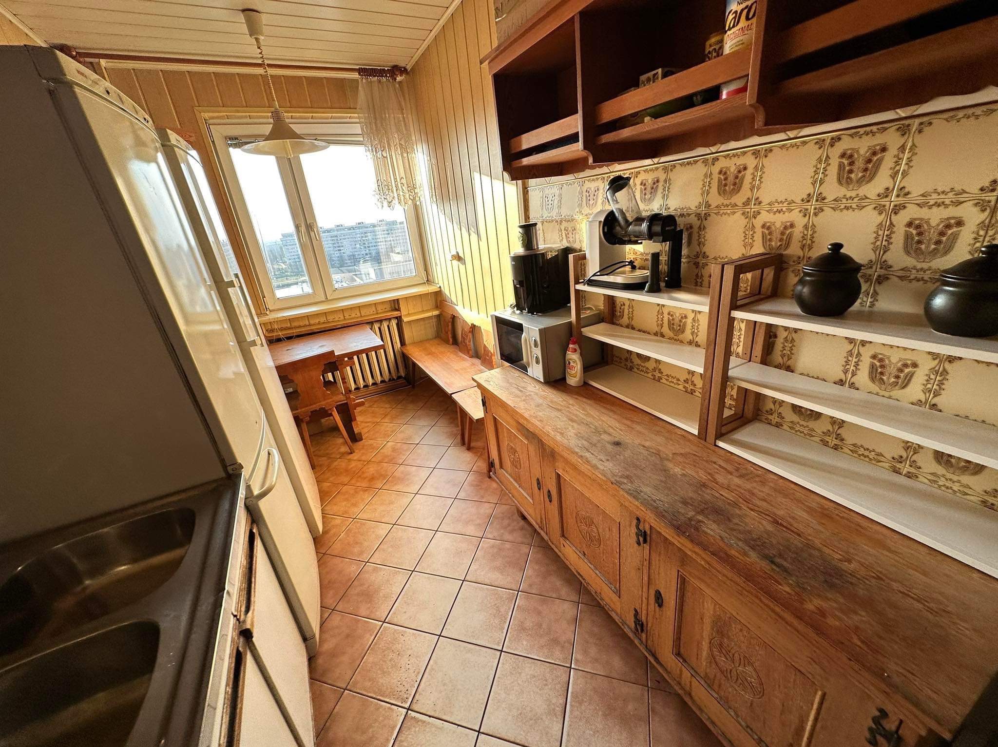 Jasna kuchnia z drewnianymi meblami, beżowymi płytkami i widokiem na miasto z okna. W tle widoczna ściana z boazerią i stylizowanymi kaflami.