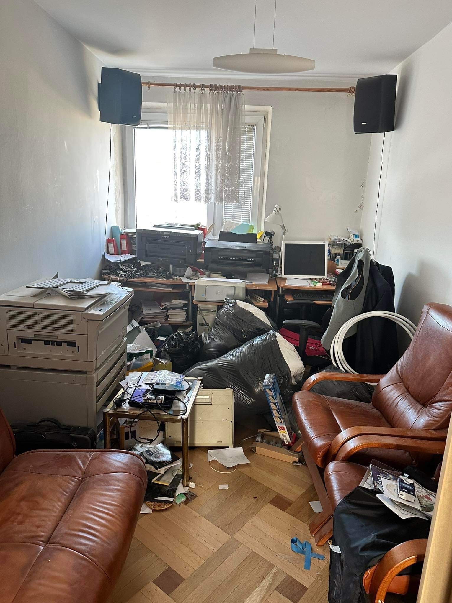 Zaniedbane biuro z brązowymi skórzanymi meblami, stertami papierów, sprzętem biurowym, workami na śmieci i głośnikami na ścianie, ukazujące potrzebę gruntownego posprzątania.