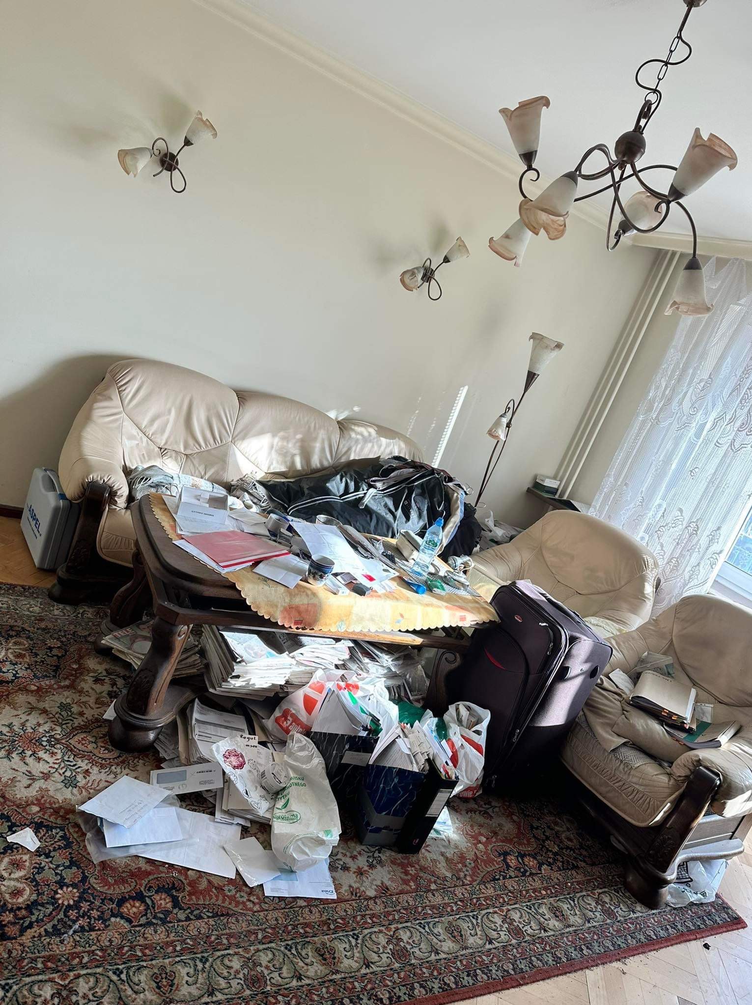 Zabałaganiony pokój dzienny z beżową kanapą, drewnianym stołem zawalonym dokumentami, walizką i dywanem w orientalne wzory przed rozpoczęciem porządkowania.