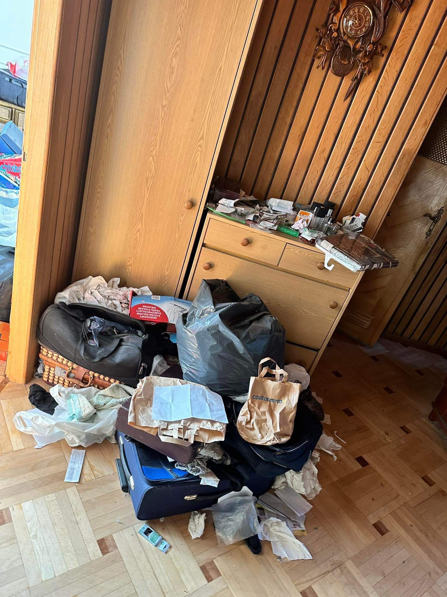 Nieuporządkowany pokój z walizkami, torbami, ubraniami i śmieciami na podłodze z parkietu, widoczne meble z drewna i zegar z kukułką na ścianie