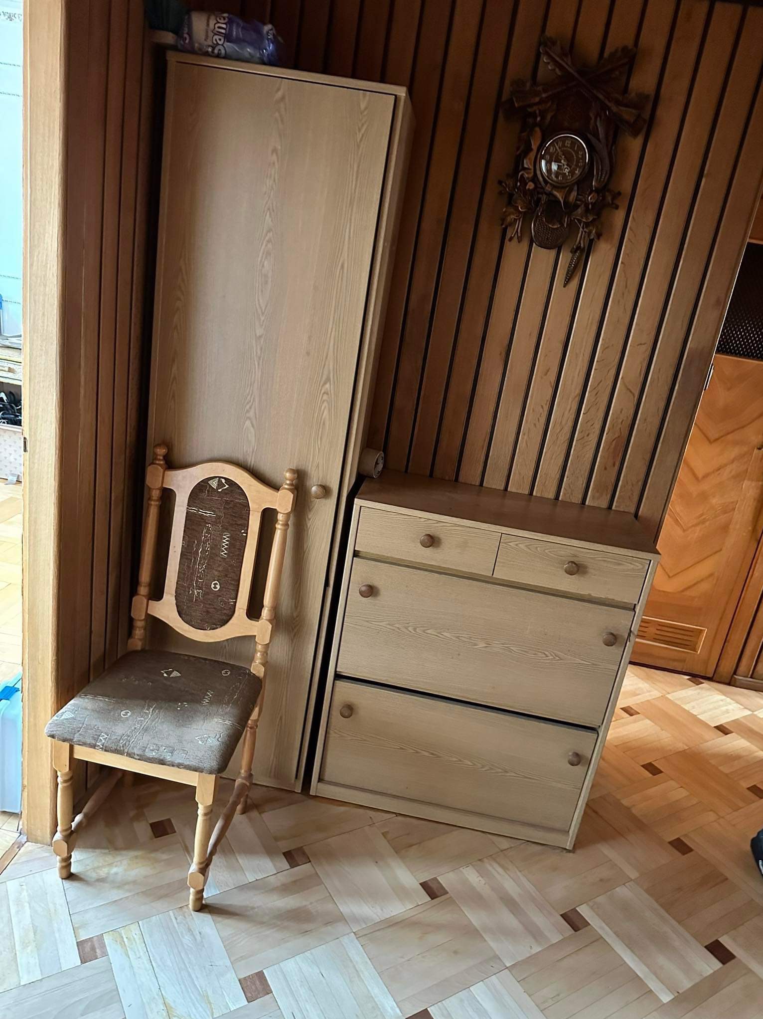 Ustawienie mebli w pokoju z boazerią: drewniana szafa, komoda z szufladami, krzesło z tapicerowanym siedziskiem i wiszący zegar z kukułką.