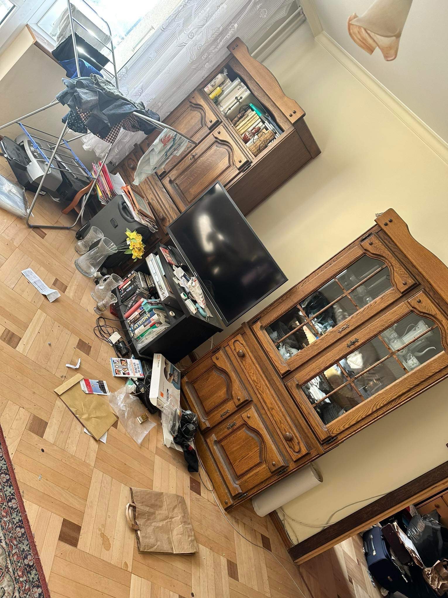 Nieporządek w pokoju: ubrania na suszarce, porozrzucane przedmioty na podłodze i półkach, widoczne meble w tle.