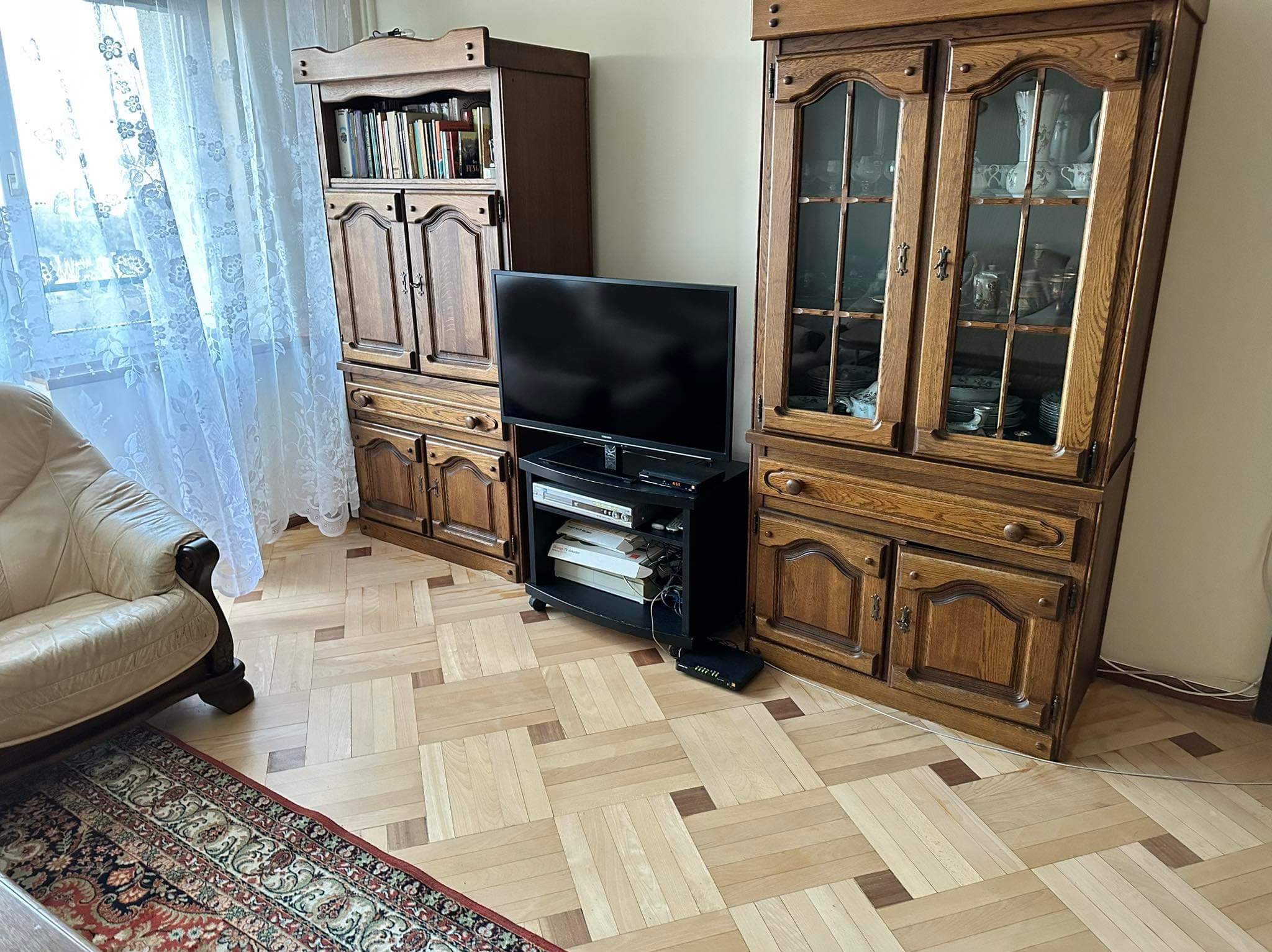 Salon z dwoma drewnianymi kredensami, czarnym stolikiem pod telewizor i wzorzystą podłogą parkietową, widok na okno z firanką.