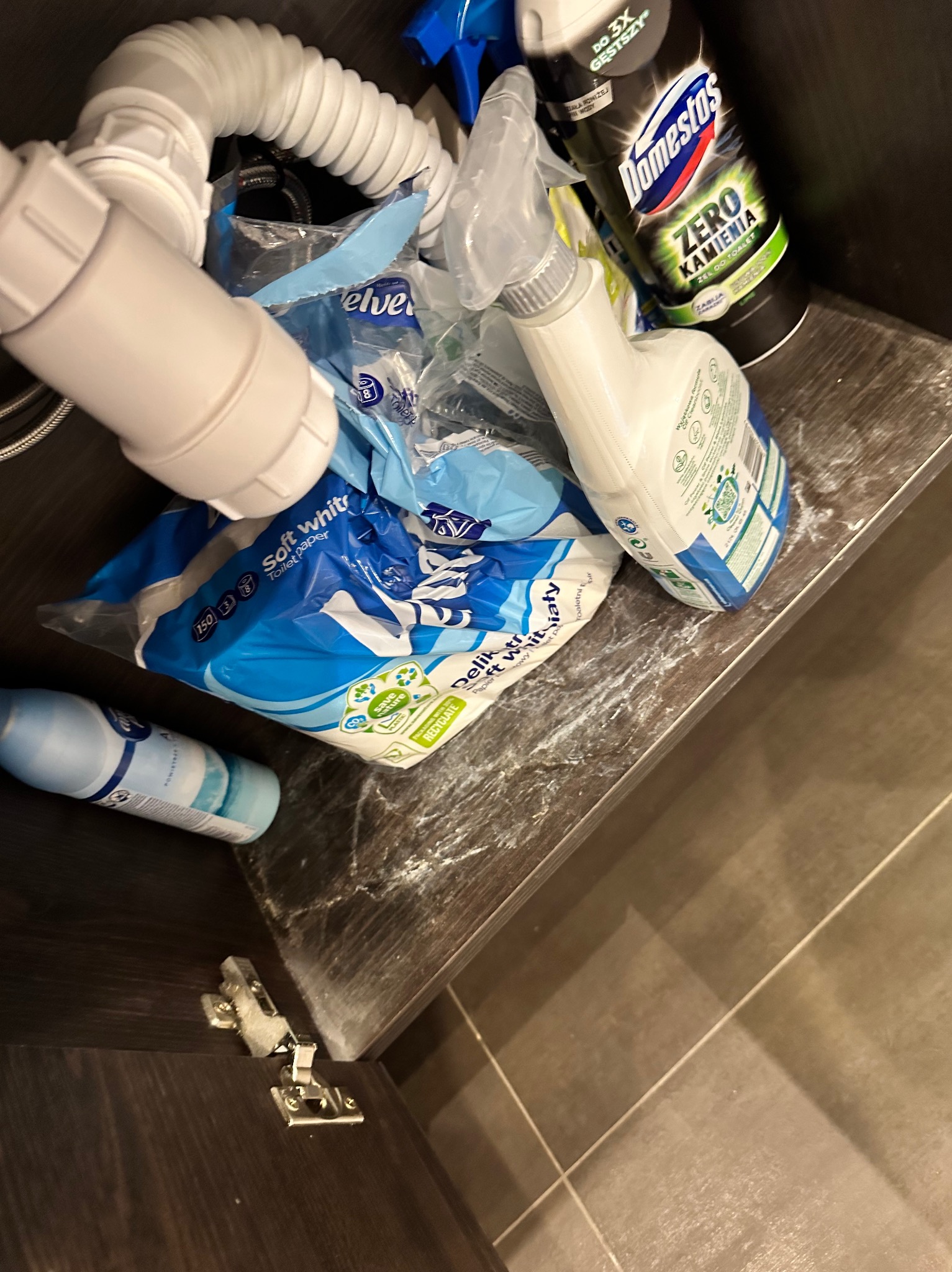 Niezorganizowana przestrzeń pod zlewem z widocznymi środkami czystości, papierem toaletowym i elementami instalacji hydraulicznej. Widoczne ślady proszku lub kurzu na blacie szafki.