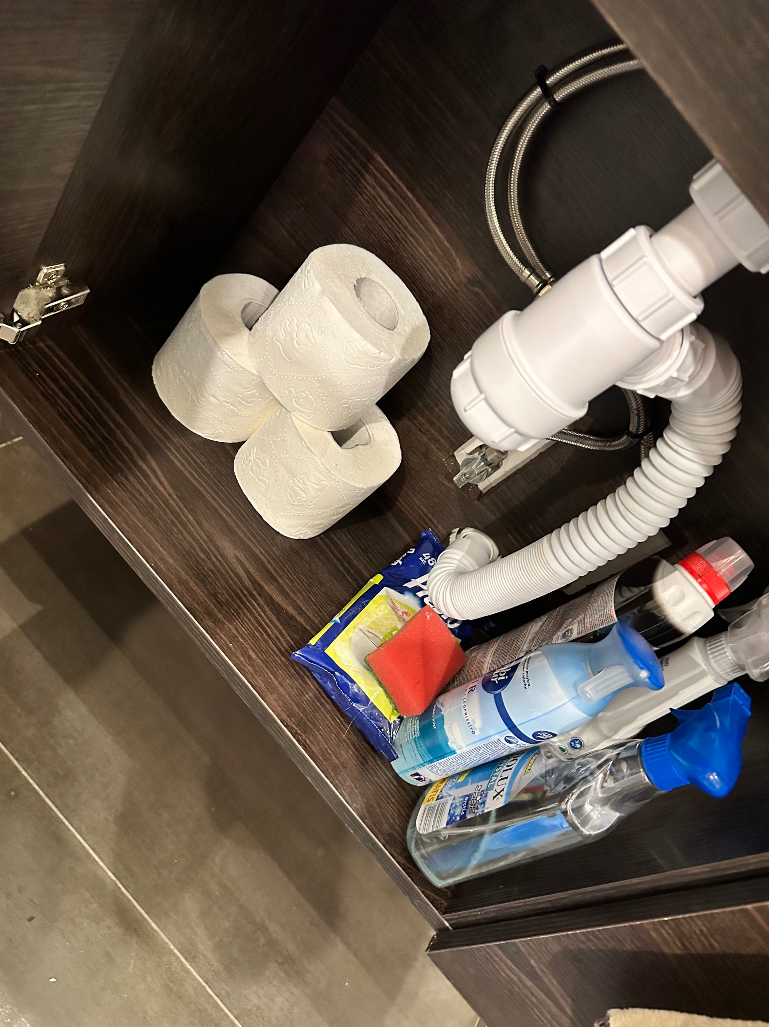 Wnętrze szafki pod umywalką z zapasem papieru toaletowego, płynami do czyszczenia w sprayu, gąbką i syfonem.