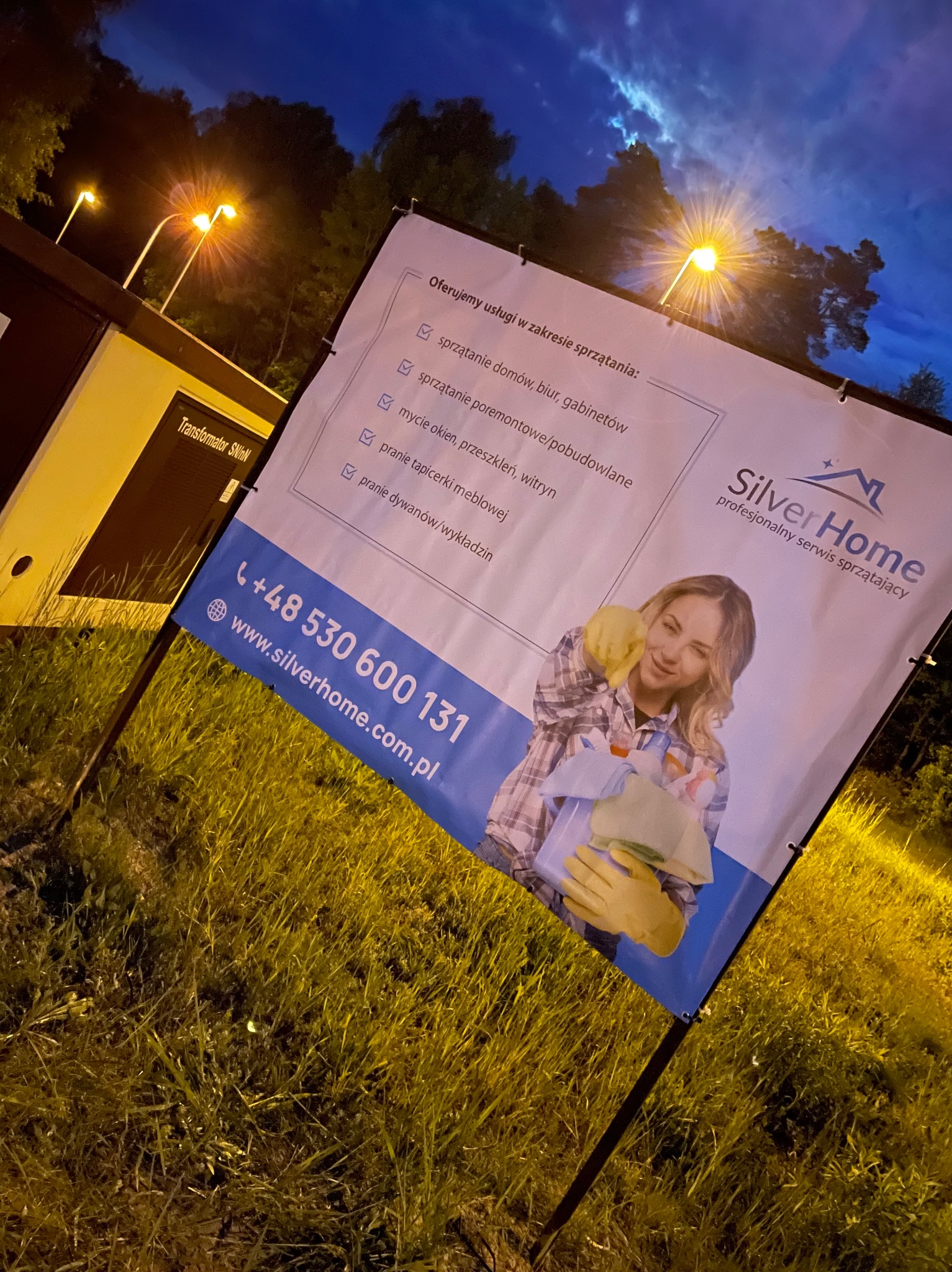 Baner reklamowy firmy Silver Home, oferującej profesjonalne usługi sprzątające, z uśmiechniętą kobietą w rękawicach trzymającą środki czystości, na tle trawy i transformatora SN/nN, oświetlony...