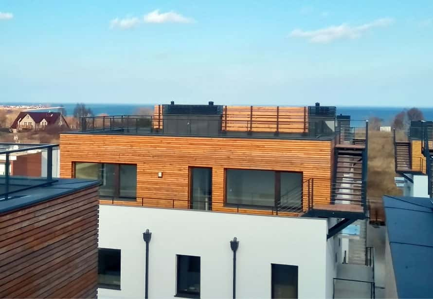 Nowoczesny apartamentowiec z drewnianą elewacją i balkonami z widokiem na morze w słoneczny dzień.