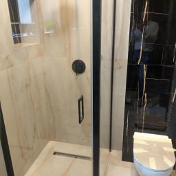 T.X.Servise - Nowoczesna kabina prysznicowa z beżowymi płytkami imitującymi marmur, czarną armaturą i odpływem liniowym w podłodze, obok toaleta zabezpieczona kartonem i taśmą, odbicie osoby w czarnej...