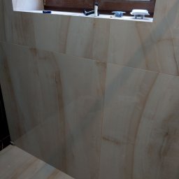 T.X.Servise - Wykończona ściana i podłoga w łazience z beżowych płytek imitujących marmur, widoczne akcesoria montażowe oświetlenia na parapecie okna.
