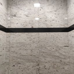 T.X.Servise - Ściana wyłożona jasnymi płytkami imitującymi kamień z ciemnymi żyłkami i czarnym, błyszczącym pasem dekoracyjnym.