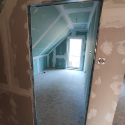 T.X.Servise - Poddasze w trakcie remontu: widok przez futrynę na pokój z oknem, ścianami z płyt gipsowo-kartonowych i częściowo nałożoną gładzią.