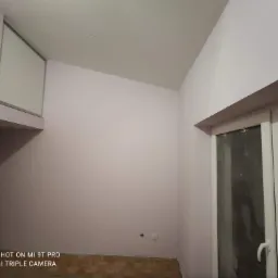 Pomalowane na jasny róż ściany w pokoju z drewnianą podłogą, widoczne okno z białymi ramami i nisko umieszczone gniazdko elektryczne.