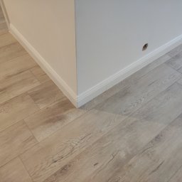 T.X.Servise - Fragment pokoju po remoncie: jasne panele podłogowe imitujące drewno, biała ściana z listwą przypodłogową i otworem po gniazdku.