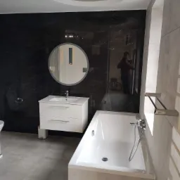 Wykończona łazienka z czarnymi marmurowymi płytkami na ścianie, białą szafką z umywalką, okrągłym lustrem z podświetleniem LED, białą wanną i szarą podłogą; widoczne odbicie osoby robiącej zdjęcie...