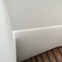 Minimalistyczne wnętrze: białe ściany, sufit z ukrytym oświetleniem LED i drewniane schody widziane z g&oacute;ry.