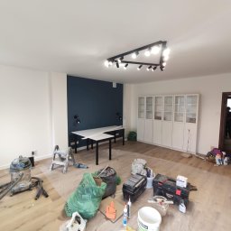 T.X.Servise - Pokój w trakcie prac wykończeniowych: zabezpieczona folią podłoga, narzędzia malarskie, odkurzacz przemysłowy, drabina malarska, ściana pomalowana częściowo na granatowo, nowoczesne oświetlenie...