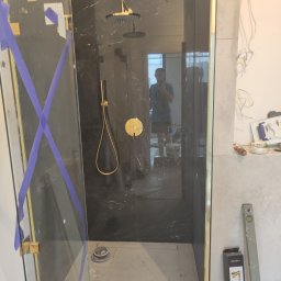 T.X.Servise - Montaż kabiny prysznicowej z czarnego marmuru i złotymi elementami wykończeniowymi, zabezpieczonej niebieską taśmą malarską, widoczne narzędzia i poziomica.