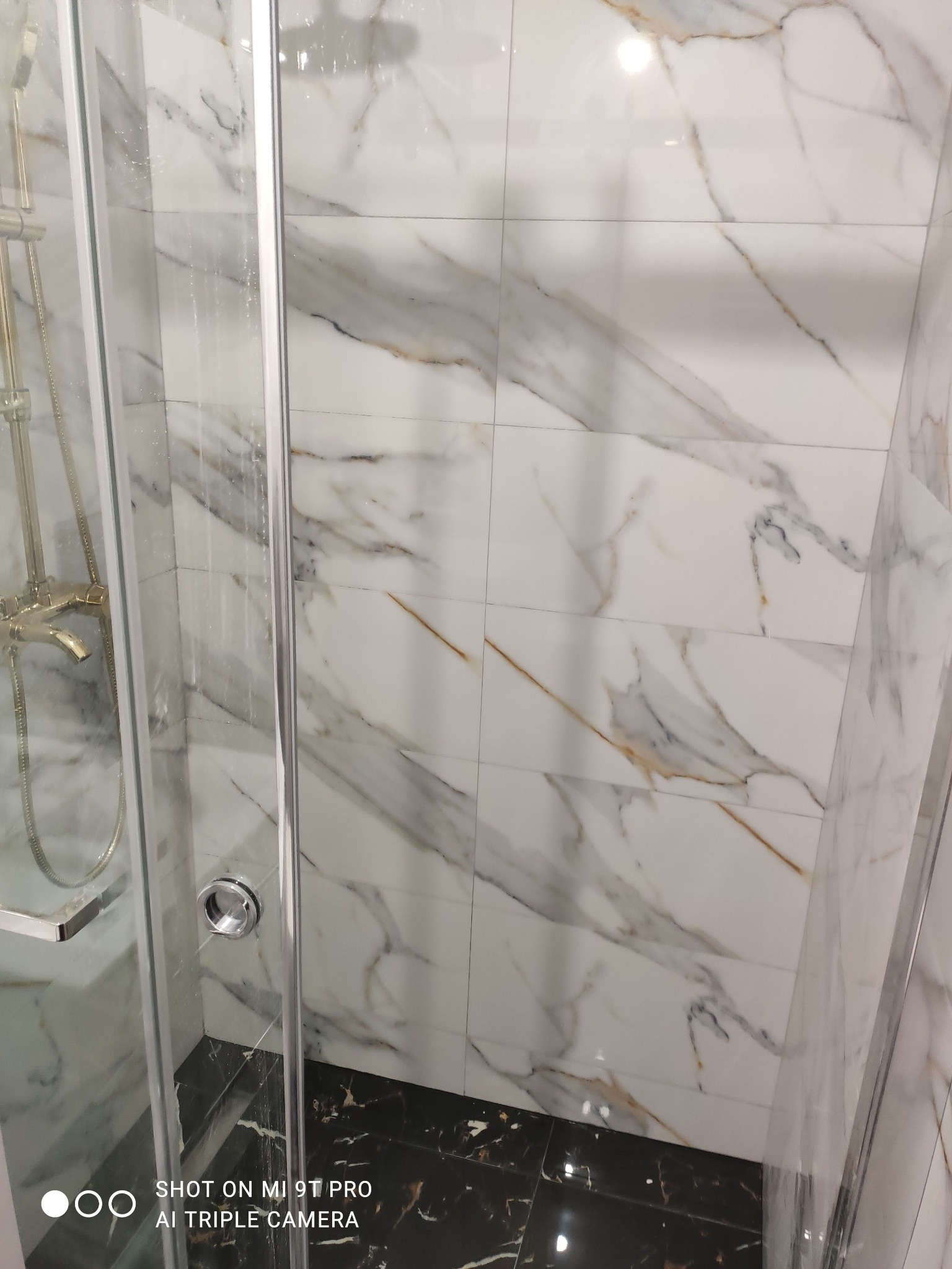Kabina prysznicowa z armaturą w kolorze złota na tle białych płytek imitujących marmur z czarną podłogą, widok z dołu.