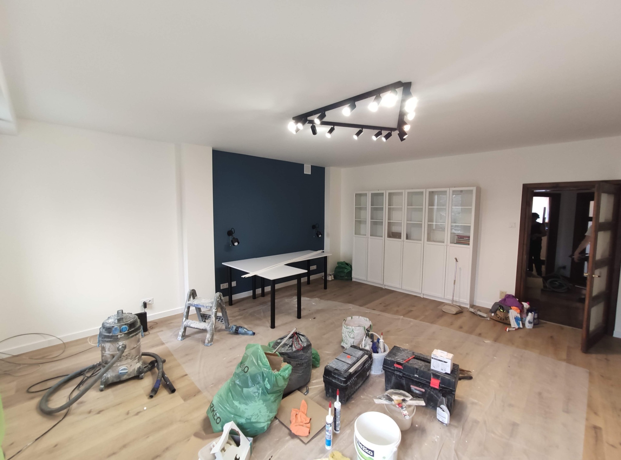 Pokój w trakcie prac wykończeniowych: zabezpieczona folią podłoga, narzędzia malarskie, odkurzacz przemysłowy, drabina malarska, ściana pomalowana częściowo na granatowo, nowoczesne oświetlenie...
