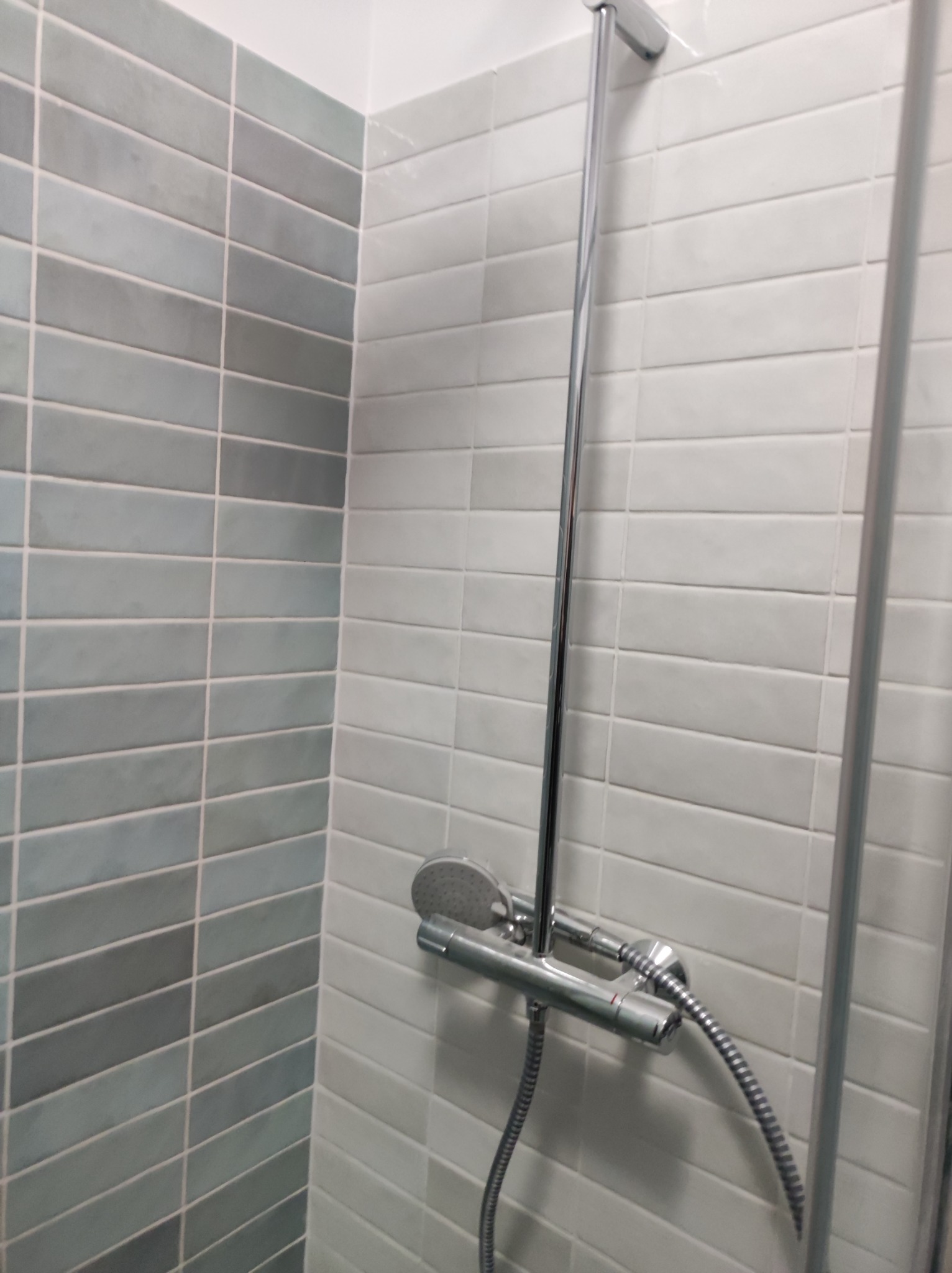 Ściana wykończona podłużnymi, szarymi i białymi płytkami w łazience, z widoczną chromowaną armaturą prysznicową.