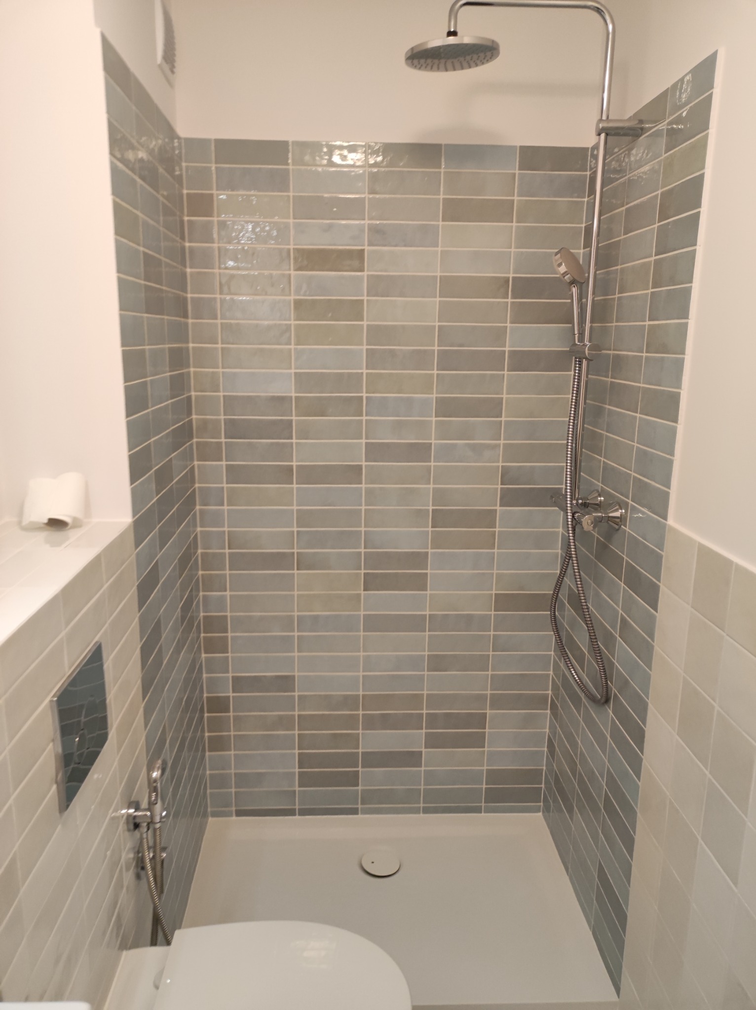 Nowoczesna kabina prysznicowa wyłożona podłużnymi, szarymi płytkami, z deszczownicą i zestawem słuchawkowym, widoczny fragment toalety z bidetką