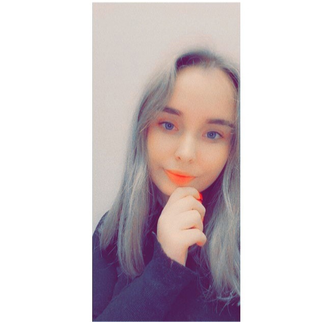 Portret młodej kobiety z popielatymi włosami, niebieskimi oczami i pomarańczową szminką, trzymającej dłoń przy brodzie.