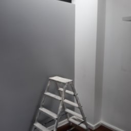 Maluj z Nami - Świeżo pomalowana szara ściana w rogu pomieszczenia z białą drabiną przystawioną do ściany, widoczna podłoga z ciemnego drewna i białe listwy przypodłogowe.