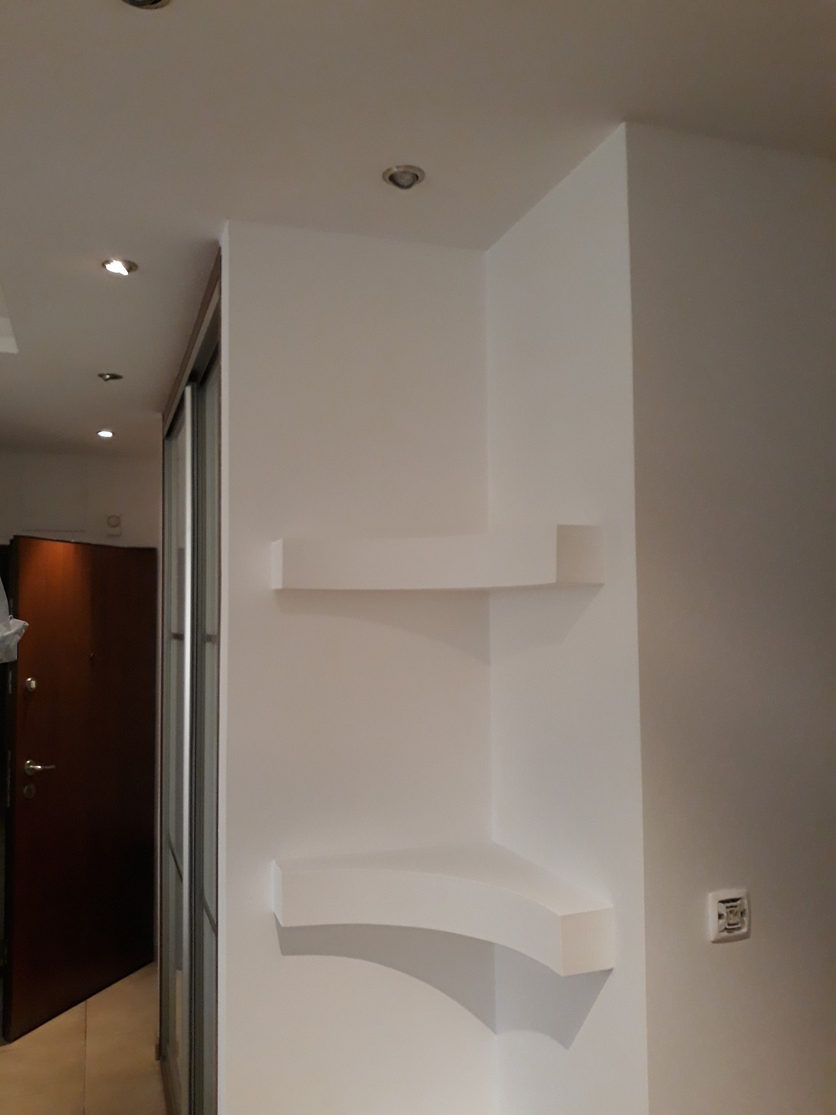 Białe, półokrągłe półki wbudowane w narożną ścianę, świeżo pomalowane na biało, z widocznym gniazdkiem elektrycznym w tle.