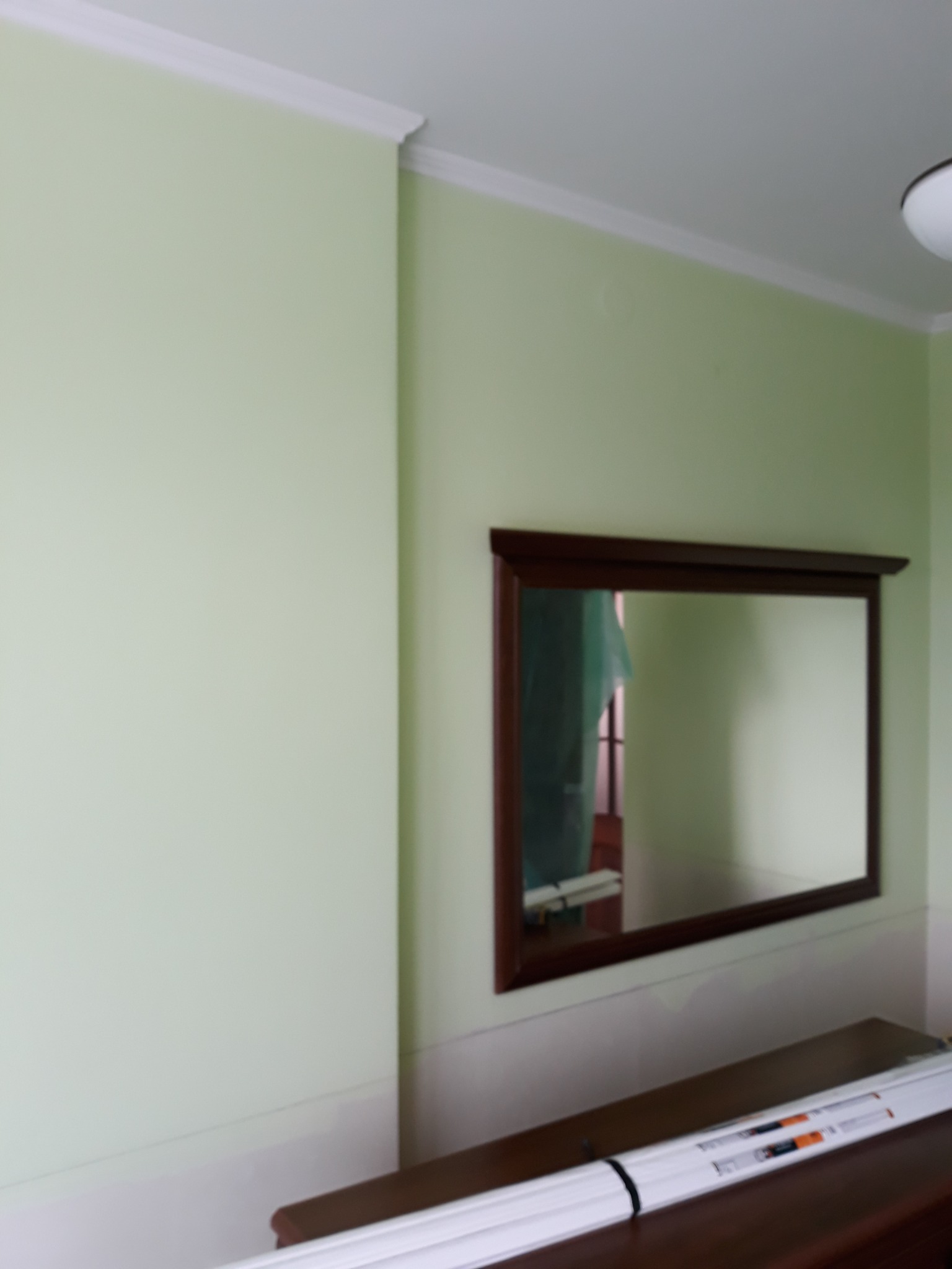 Jasnozielona ściana z brązową ramą lustra, białe listwy przypodłogowe i sufitowe, oraz zapakowane lampy LED na ciemnobrązowej komodzie.