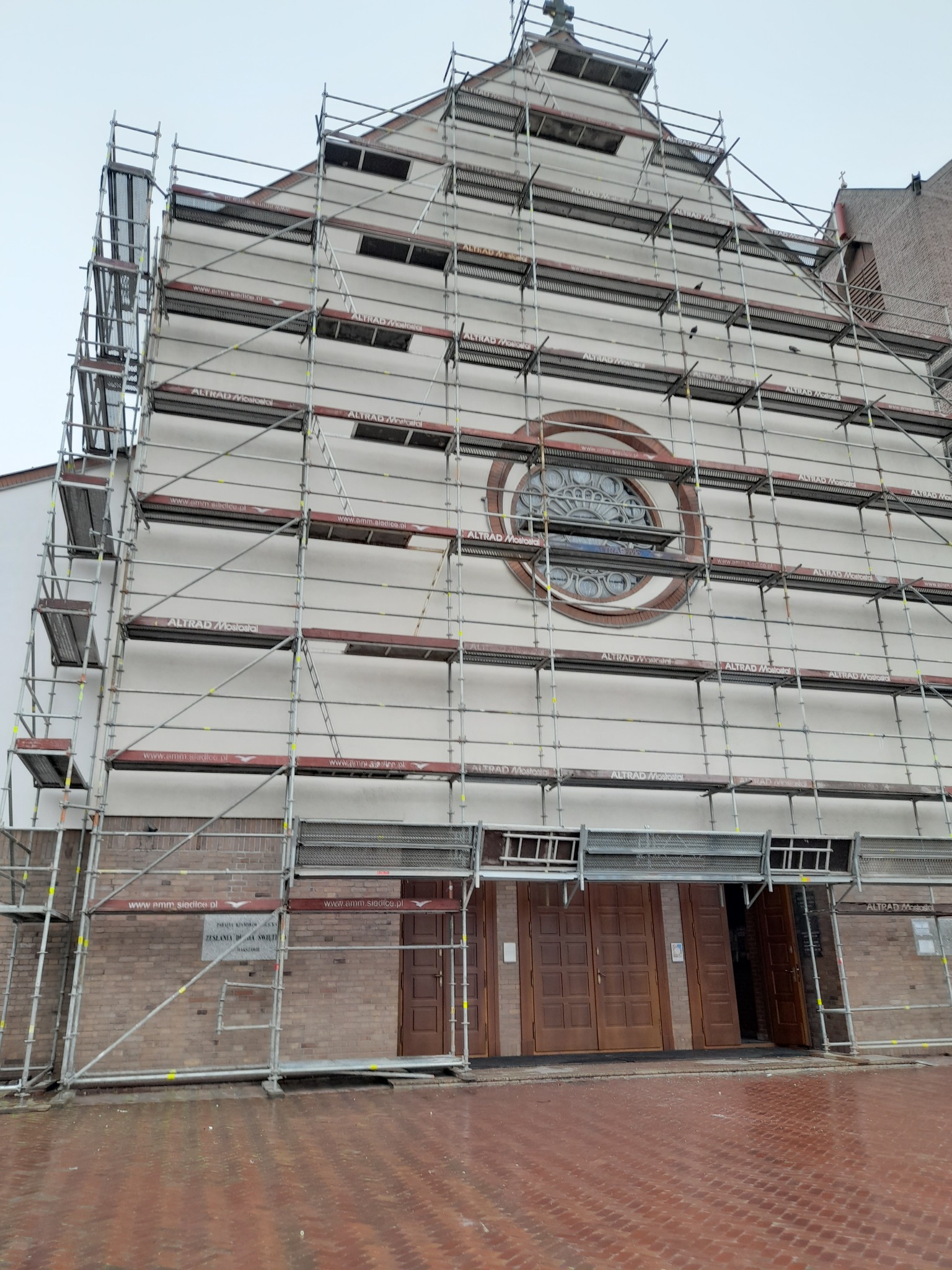 Kościół w trakcie renowacji elewacji, otoczony rusztowaniami firmy Altrad Mostostal, z widocznym okrągłym oknem i ceglanym wejściem.