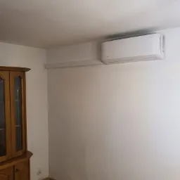 Biała jednostka klimatyzacji zamontowana na ścianie w pomieszczeniu, widoczna instalacja elektryczna w listwie, obok stoi drewniany kredens.
