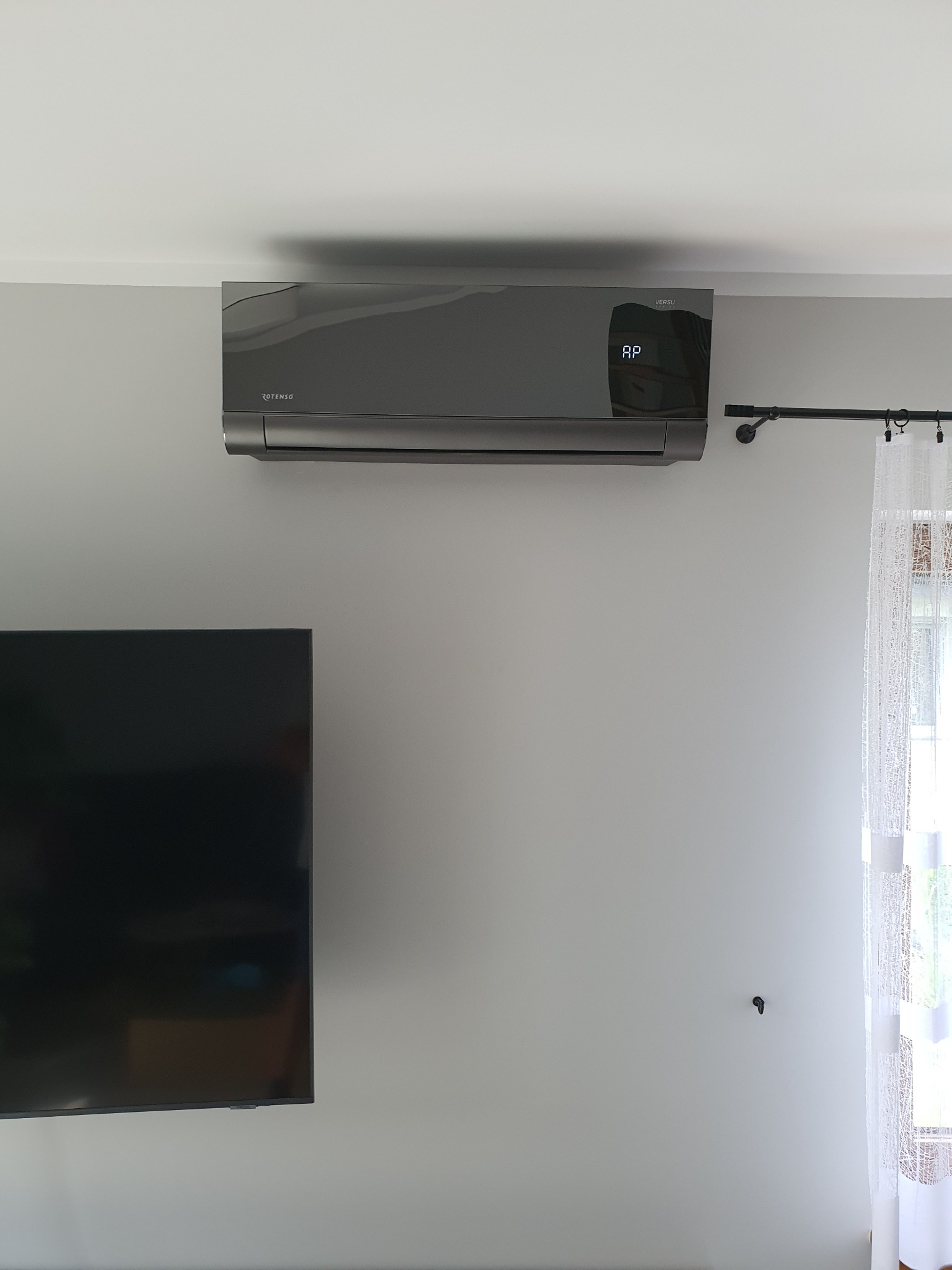 Szary klimatyzator marki Rotenso Versu zamontowany na ścianie w jasnym pomieszczeniu, widoczny fragment telewizora i zasłony.