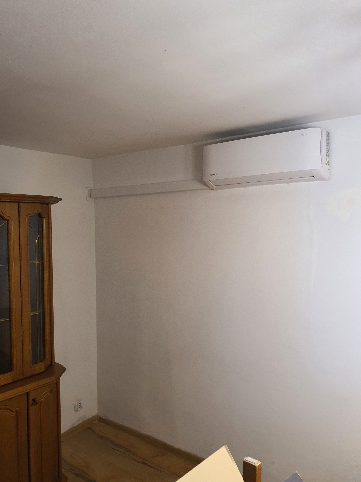 Biała jednostka klimatyzacji zamontowana na ścianie w pomieszczeniu, widoczna instalacja elektryczna w listwie, obok stoi drewniany kredens.