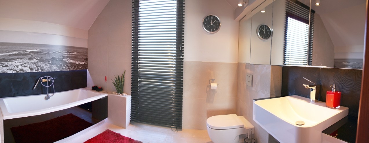 Nowoczesna łazienka na poddaszu z wanną, umywalką, toaletą, lustrem w szafce, roletami okiennymi i fototapetą z motywem morza.