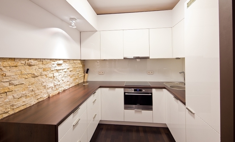 Nowoczesna kuchnia z białymi, lakierowanymi szafkami, drewnianym blatem i ścianą wyłożoną dekoracyjnym kamieniem piaskowcem. W tle widoczny piekarnik elektryczny i płyta indukcyjna.