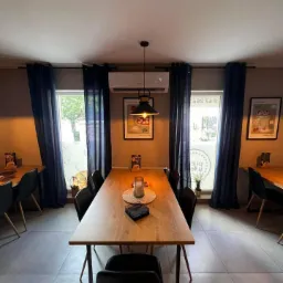 Wnętrze restauracji z długim drewnianym stołem, czarnymi krzesłami, zasłonami w oknach i zamontowanym klimatyzatorem.