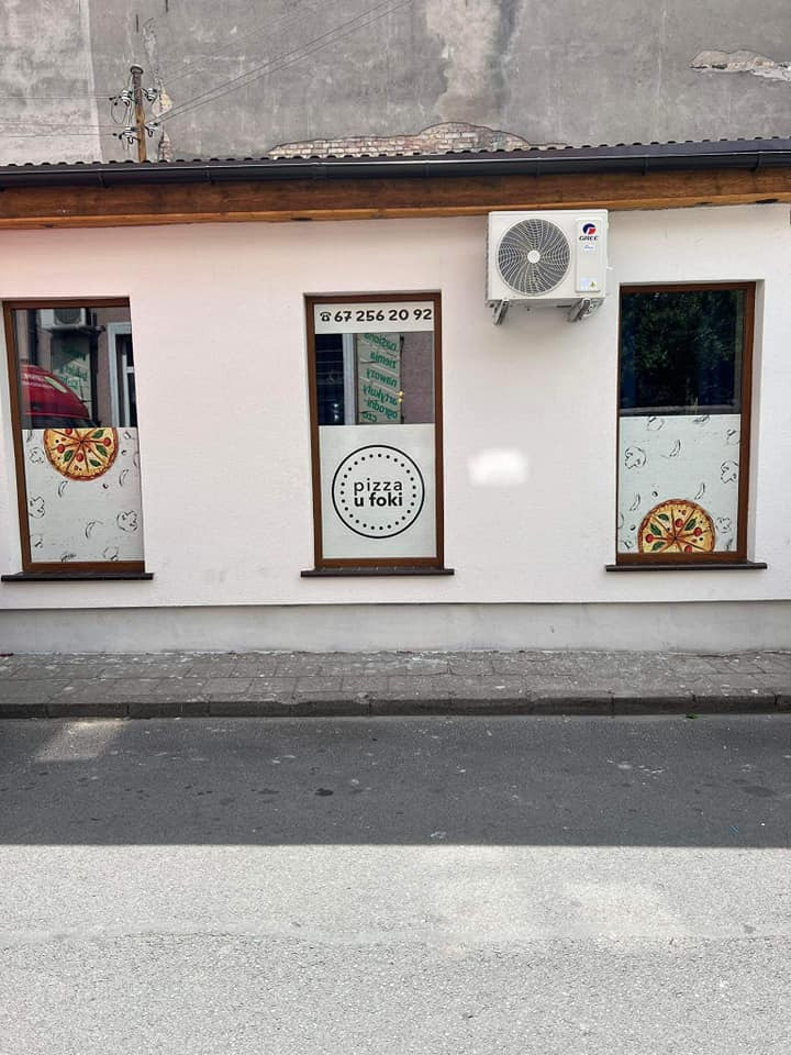 Biała fasada pizzerii z trzema oknami, na środkowym numer telefonu i logo, na skrajnych naklejki z pizzą, a nad jednym z okien zamontowany klimatyzator.
